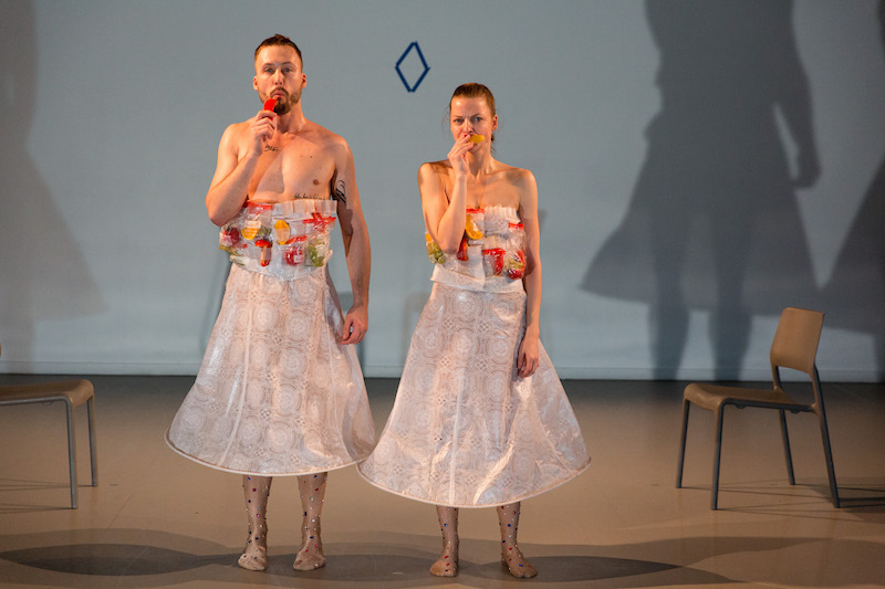 Martin and Denisa wear white flowered plastic looking long skirts and jeweled tights. They look like they're in mid chew of eating something